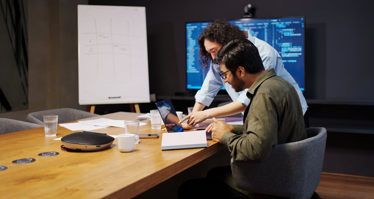Dos personas revisando prototipos y bocetos en una mesa de trabajo con laptop y monitor